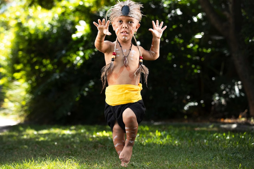 A young indigenous boy with a form of dwarfism dances in traditional dress. He looks to the camera with his palms spread.