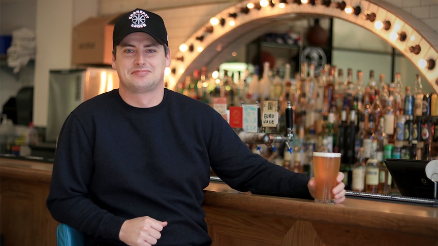 Lachlan Stevens sits at a bar wearing a black shirt and hat while holding a beer.