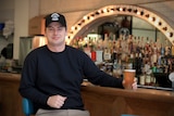 Lachlan Stevens sits at a bar wearing a black shirt and hat while holding a beer.