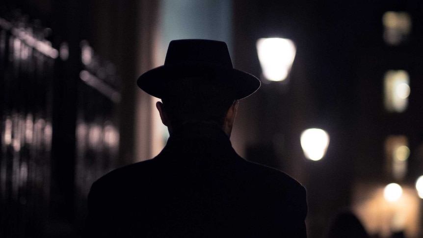 A dark street showing the back of a man's head, wearing a hat, reminiscent of noir film streetscapes