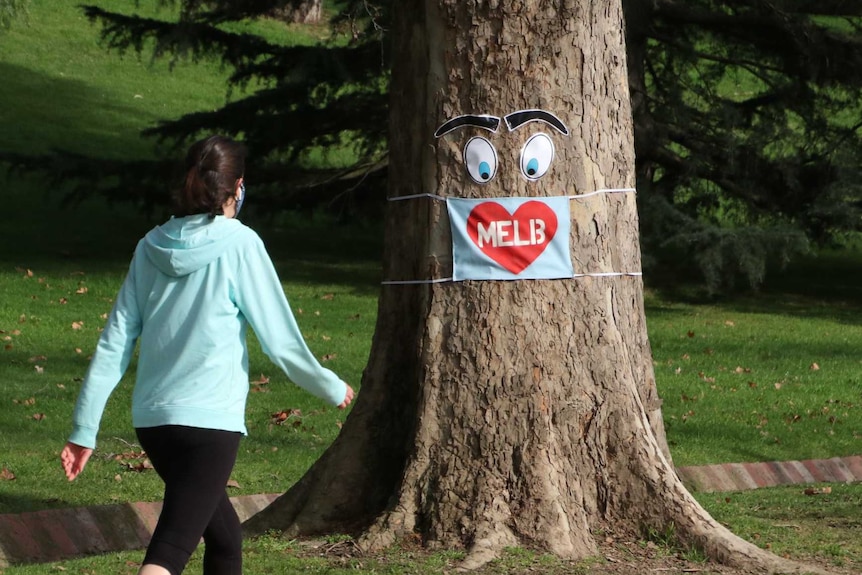A woman walks towards a tree with a pair of funny eyes and a Melb facemask on it.