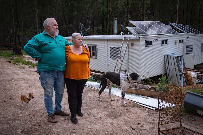 A couple stand with arms around each other, with their two dogs in front of a metal building.