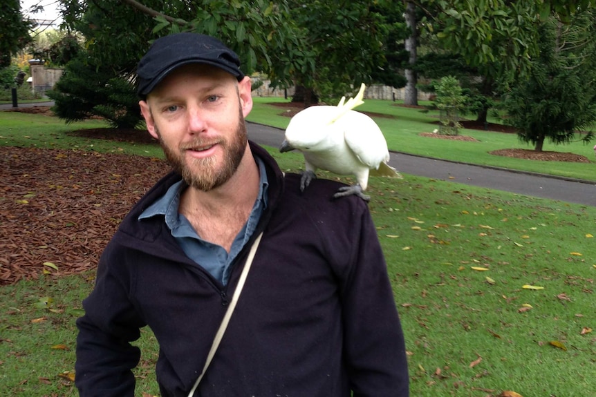 Dr Martin smiling with a cockatoo in a park, the cockatoo appear to be looking at him, there is an ibis in the background