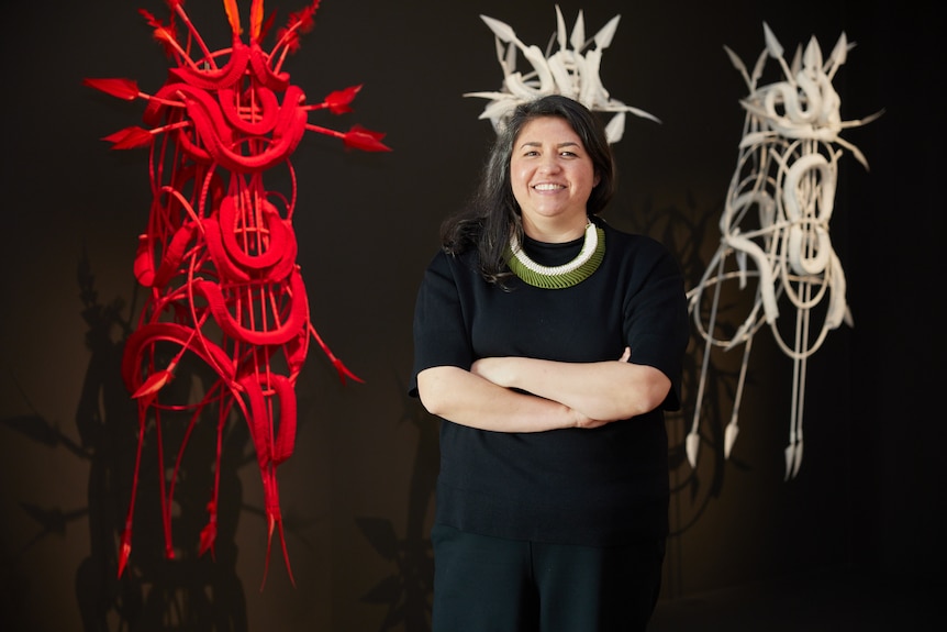 A First Nations woman in black dress and green and white necklace stands in front of three elaborate sculptures