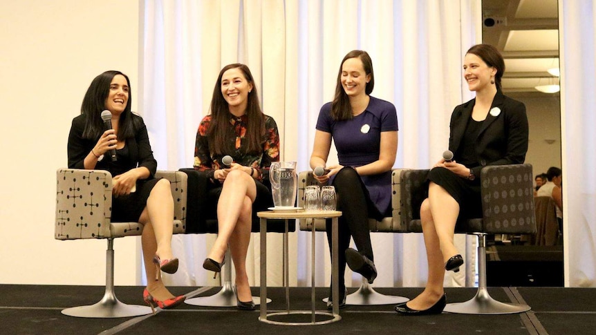 Four young women laugh while holding microphones, sitting on a stage in formal clothing.