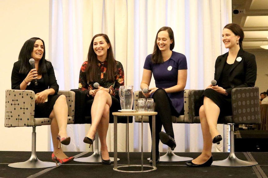 Four young women laugh while holding microphones, sitting on a stage in formal clothing.