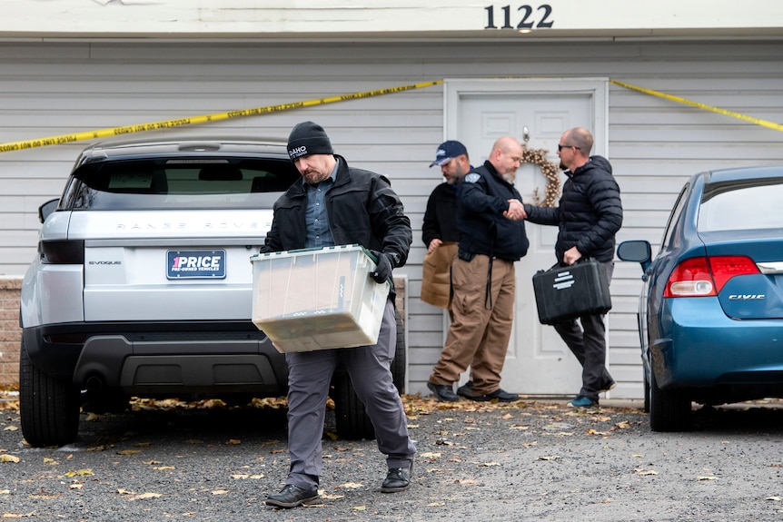 A man wearing a beanie removes a plastic box from a house cordoned off with police tape