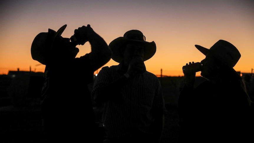 Three people wearing hats, two drinking from cans, are silhouetted as the sun sets.