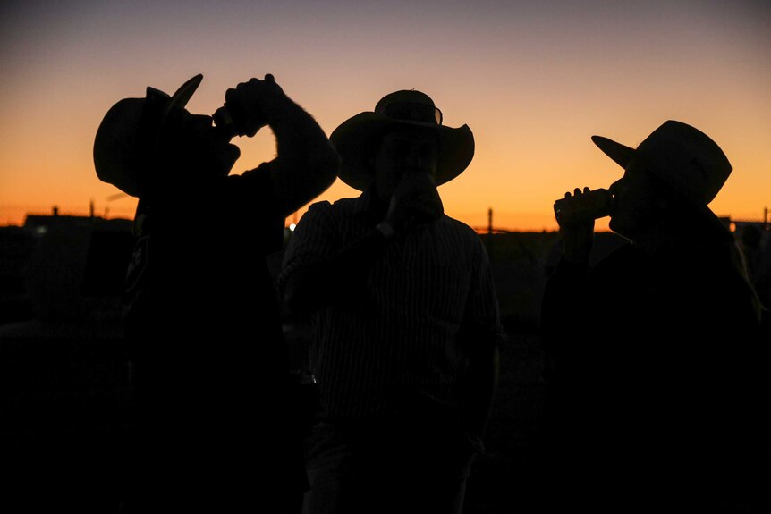 Three people wearing hats, two drinking from cans, are silhouetted as the sun sets.