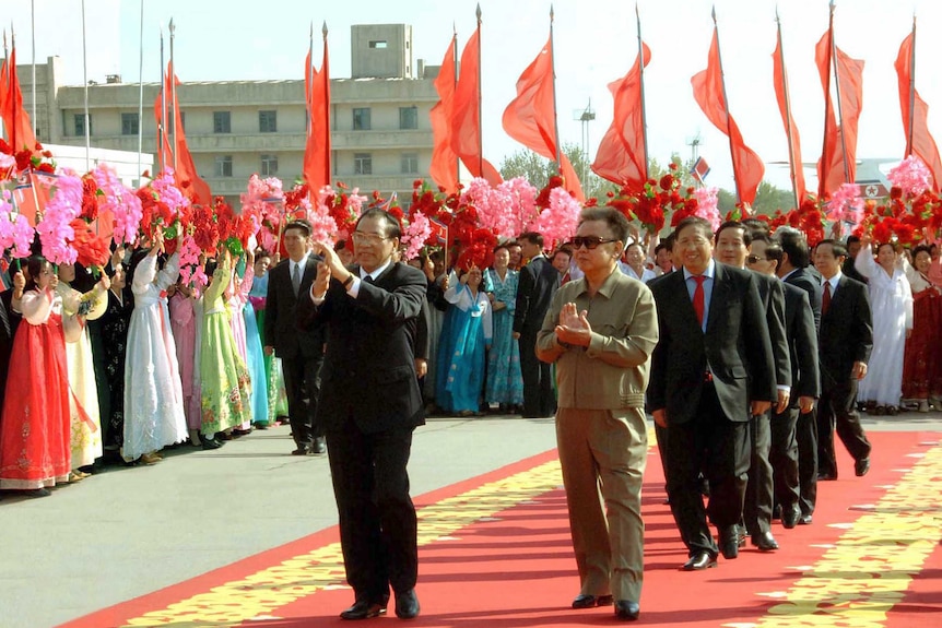 Outside two leaders walk on a red carpet as women in bright North Korean dress wave pink flowers and red flags flutter behind.