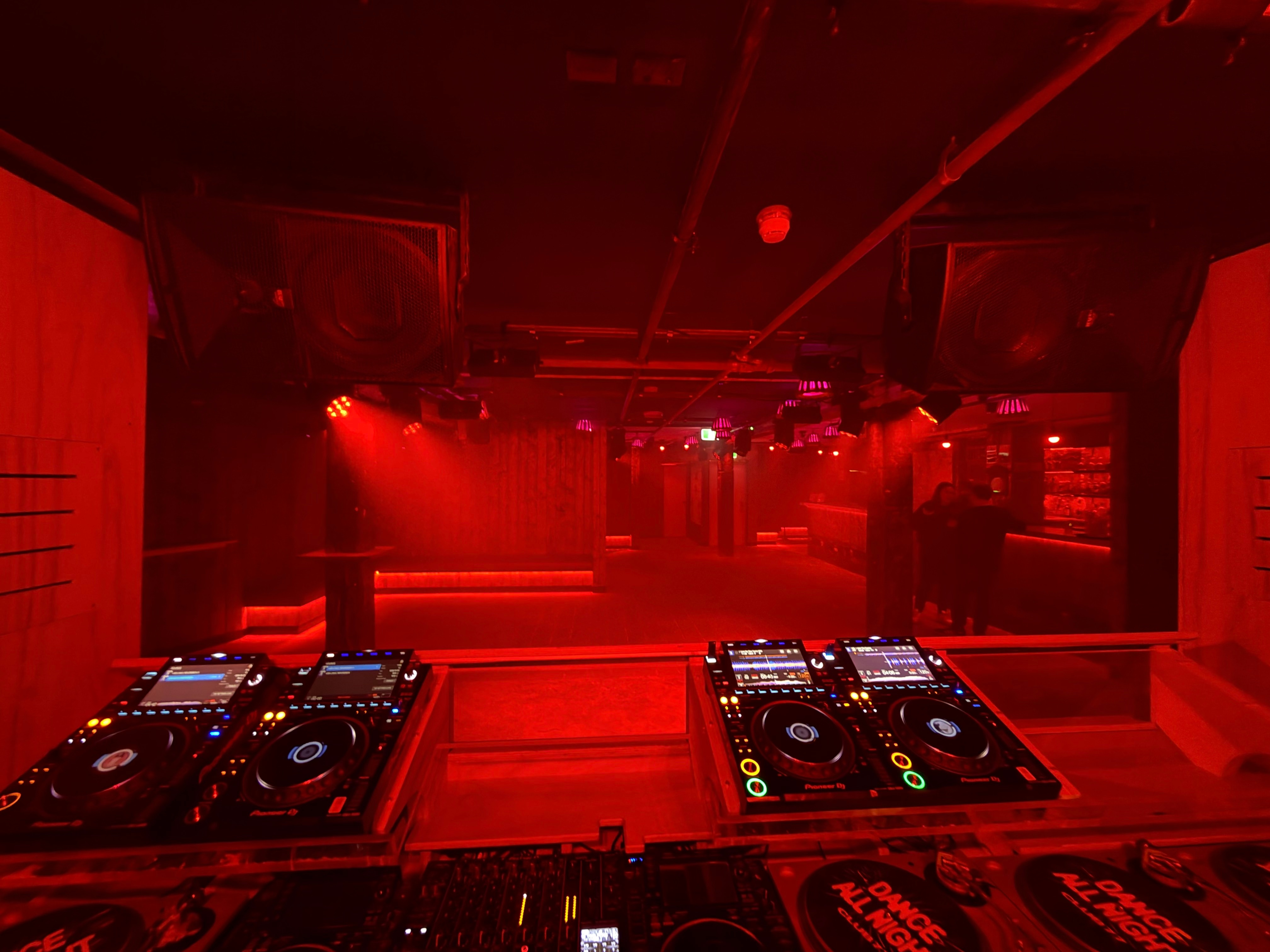A red lit room with DJ decks in the centre, and an empty dancefloor