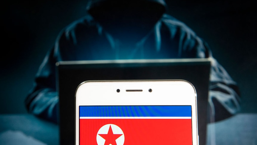 A shadowy hooded hacker figure behind a laptop with a phone with the North Korean flag in front