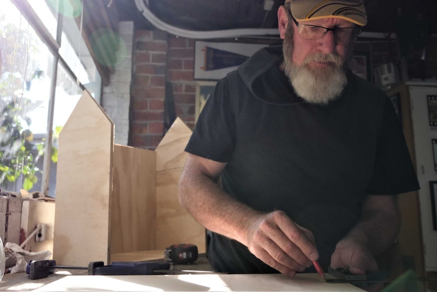 Man ruling a line on a piece of timber on a bench, with a half built tiny timber house behind him.