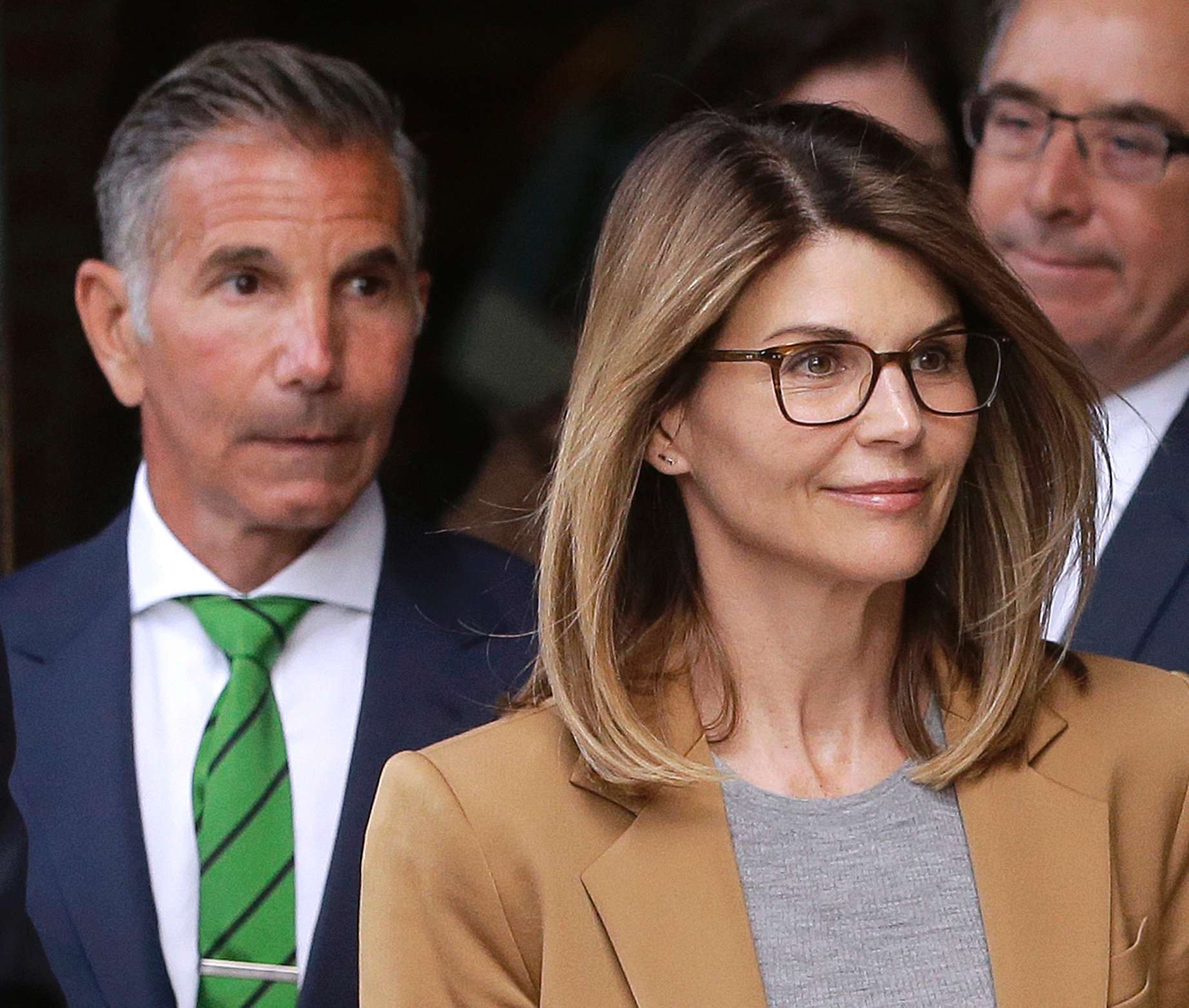 Lori Loughlin wears a grey sweater and tan coat and glasses and there are several men in suits behind her