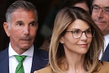 Lori Loughlin wears a grey sweater and tan coat and glasses and there are several men in suits behind her