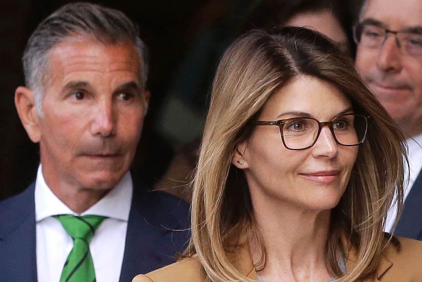 Lori Loughlin wears a grey sweater and tan coat and glasses and there are several men in suits behind her