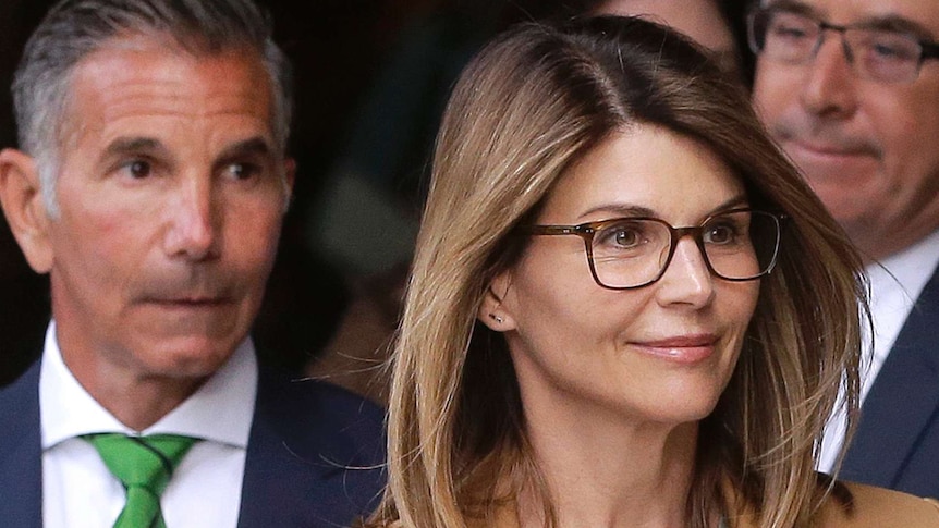 Lori Loughlin wears a grey sweater and tan coat and glasses and there are several men in suits behind her