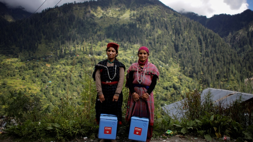 Two women stand together in front of a green mountain backdrop, holding a big blue container each.