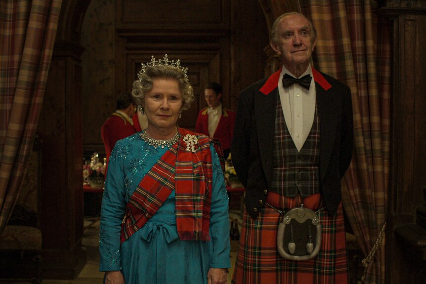 Queen Elizabeth and Prince Philip enter an event. Elizabeth wears a blue dress and a sash made of checked Scottish cloth.