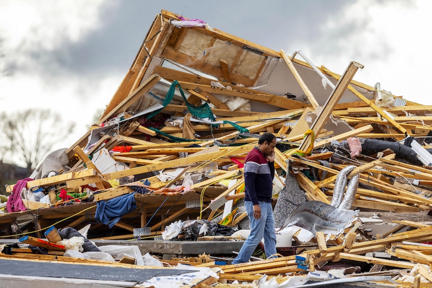A man walking past a house destroyed into a pile of debris. 
