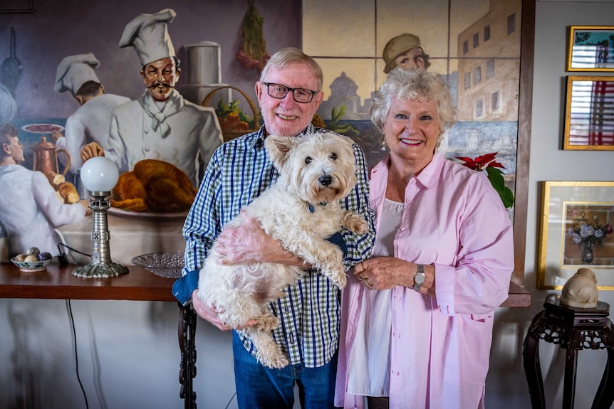 John and Bev stand in front of a colourful painting while holding a small dog.