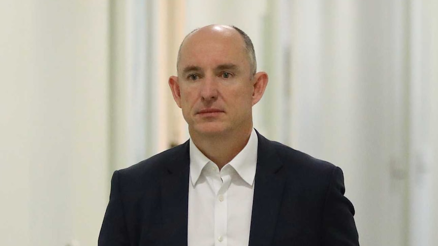 A man wearing a dark suit with a white shirt walks down a hallway, carrying an iPad.