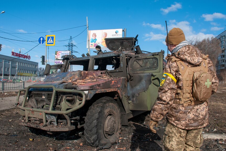 A Ukrainian soldier inspects a damaged vehicle with a flat tire and burn marks.