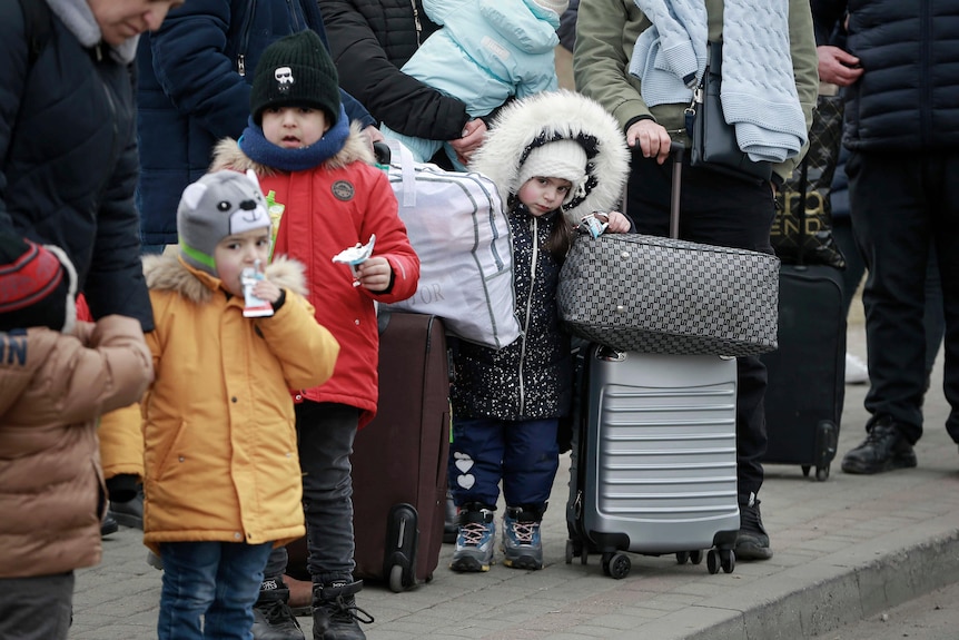 Young children stand in a line with their parents and suitcases wearing beanies and thick winter coats.