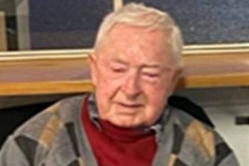An elderly man wearing a jacket sits on a chair.
