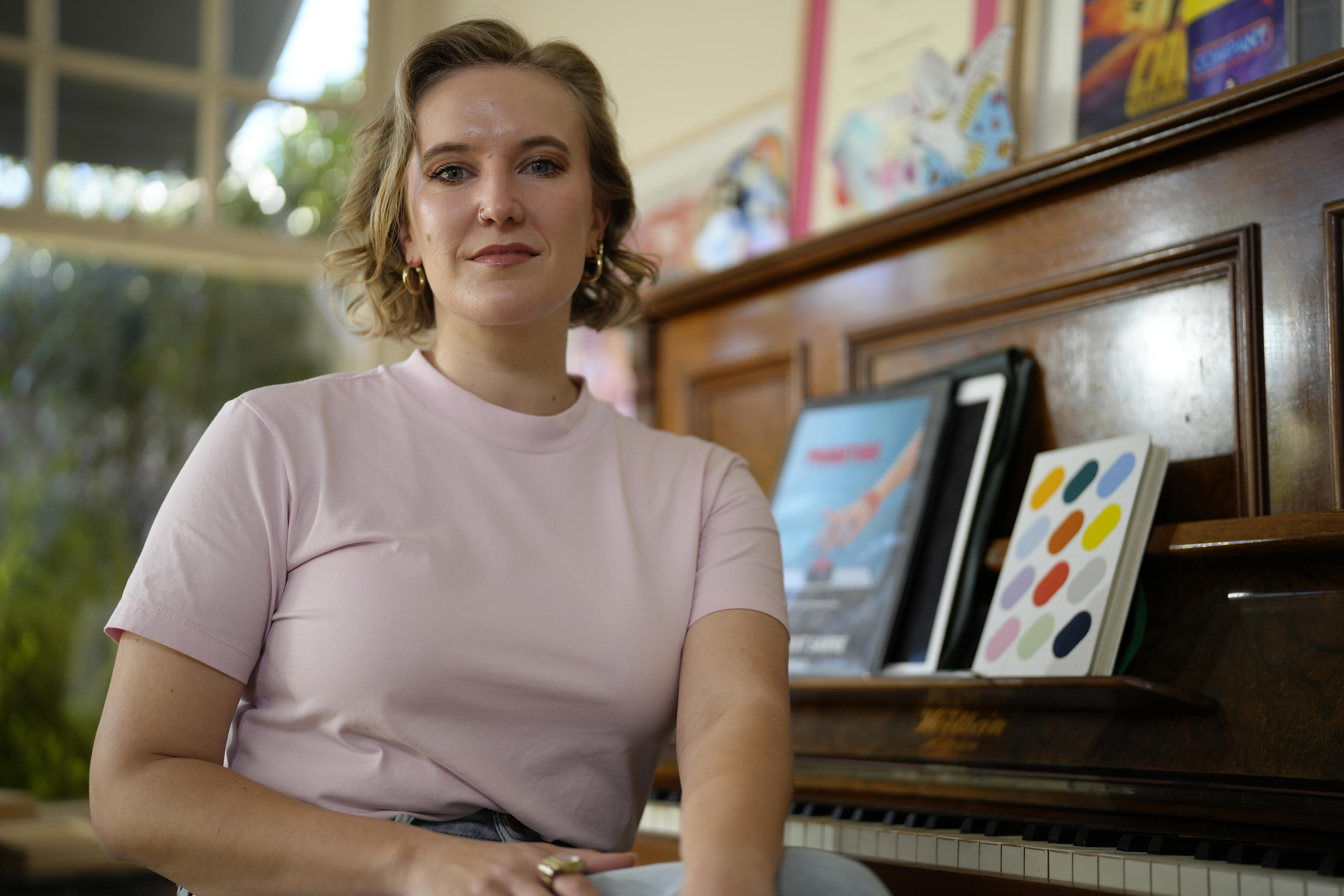 Millicent Sarre looks at the camera. She wears a pink t-shirt and hoop earrings. A brown piano with books is in the background.