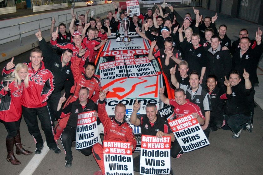 People crowd around a Holden car holding up their pointer finger in a "number one" sign.