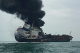 A large vessel tilts on its side in the water as thick black smoke billows rises from it into the sky.