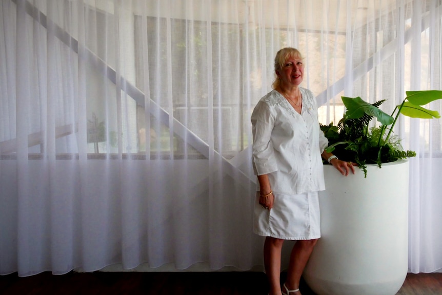 A woman in a white shirt and skirt with white-gray hair stands by a pot plant in a restaurant.