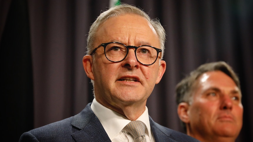 Albanese, wearing black rimmed glasses, stands in front of blue curtain speaking.