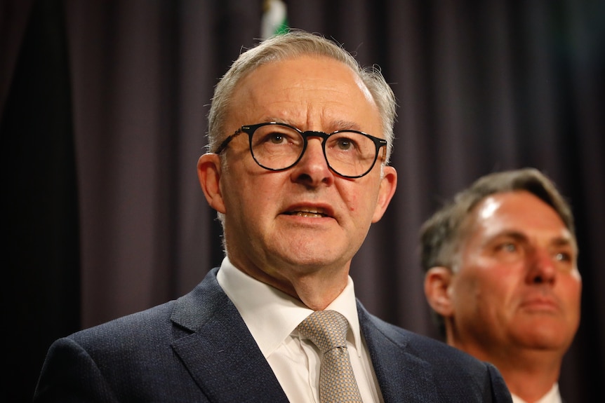 Albanese, wearing black rimmed glasses, stands in front of blue curtain speaking.