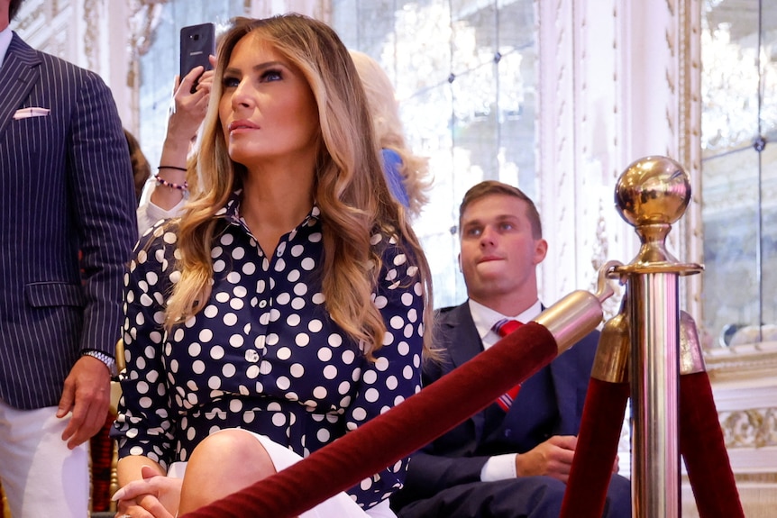 Melania Trump sits on a chair among a crowd of people behind ropes.