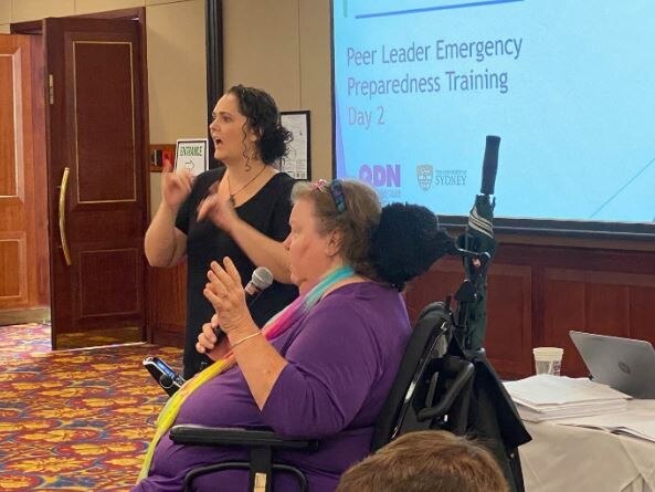 Two women at the front of a room talking into microphones. One woman is in a wheelchair.