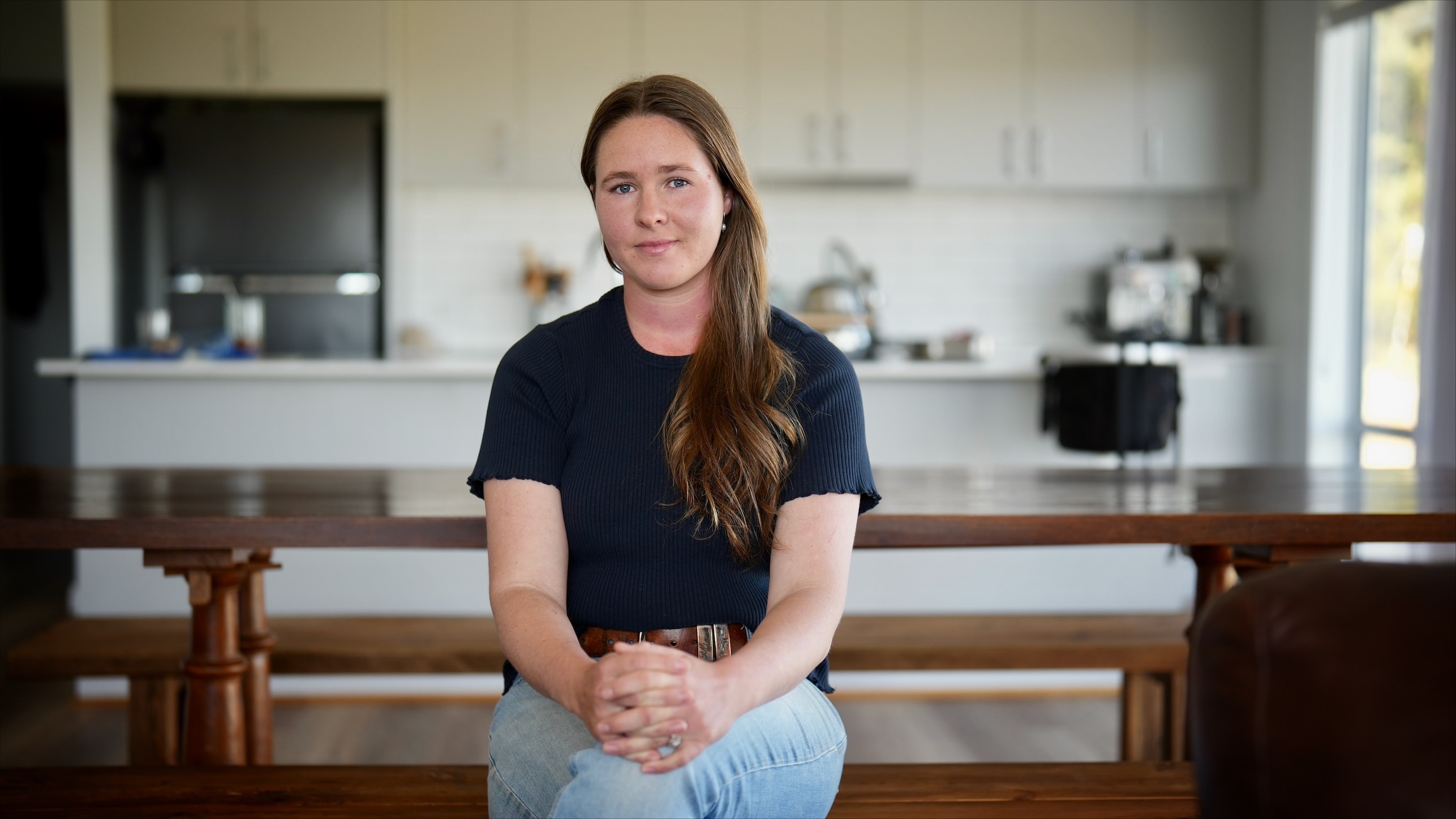 A long-haired woman (Shara Kimpton) sits cross-legged at at dining table in her kitchen.