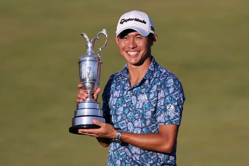 A smiling, cap-wearing golfer holds The Open trophy in his hands to his side after winning the tournament.