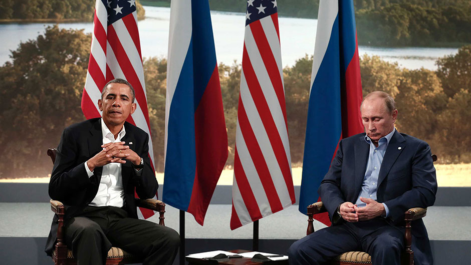 Barack Obama and Vladimir Putin looking glum while sitting in chairs before a row of US and Russian flags