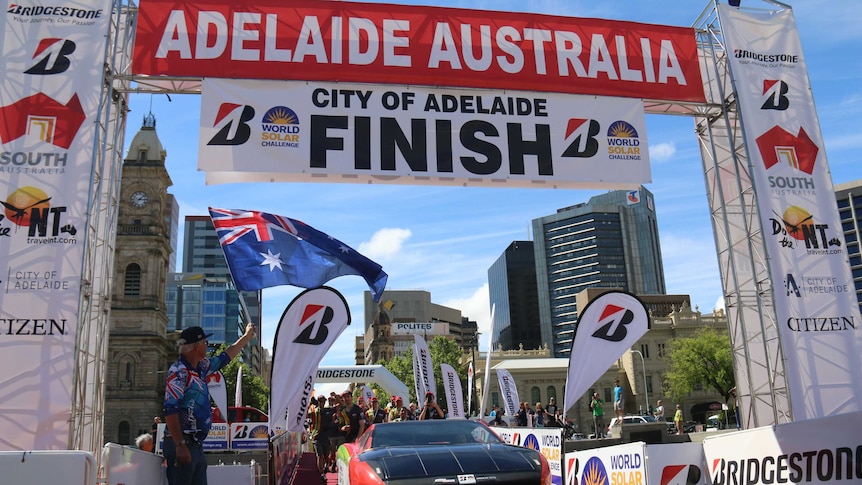 A group of people cheer under the finish line next to their team car.