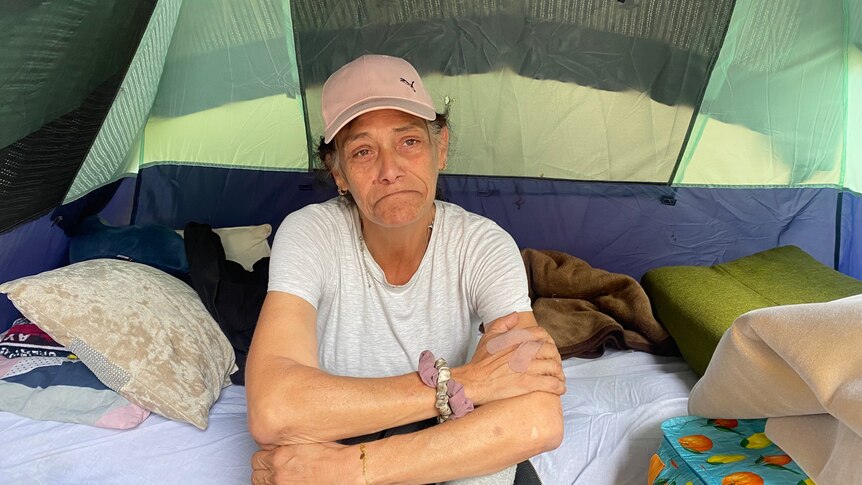 An image of a woman, frowning, sitting in a tent with her belongings scattered around her
