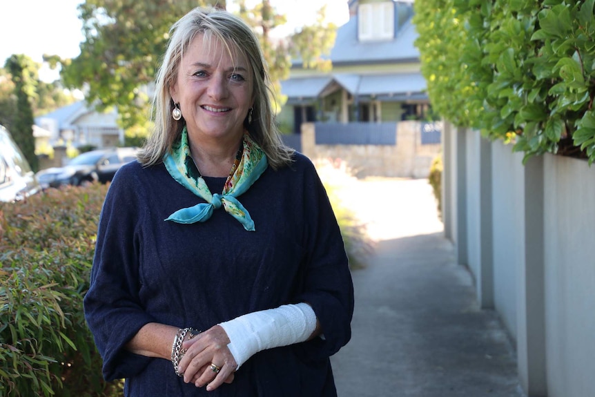 A woman in a blue top stands on a suburban street with her arm bandaged.