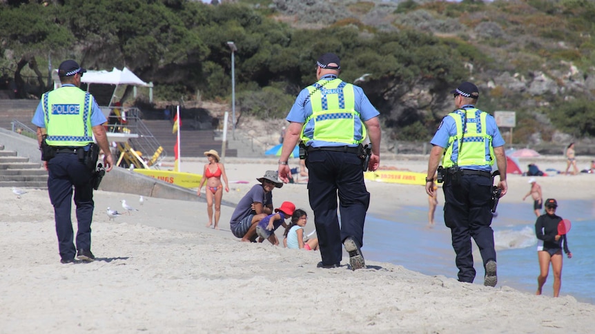 Three police officers walk along Cottesloe Beach in uniform with beachgoers nearby on the sand.