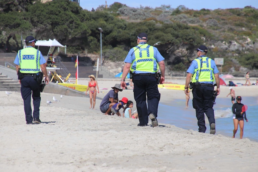 Three police officers walk along Cottesloe Beach in uniform with beachgoers nearby on the sand.