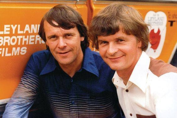 Old colour photo of two smiling men in 70s hairstyle and clothes. Both wear shirts, in front of Leyland Brothers Films sign.