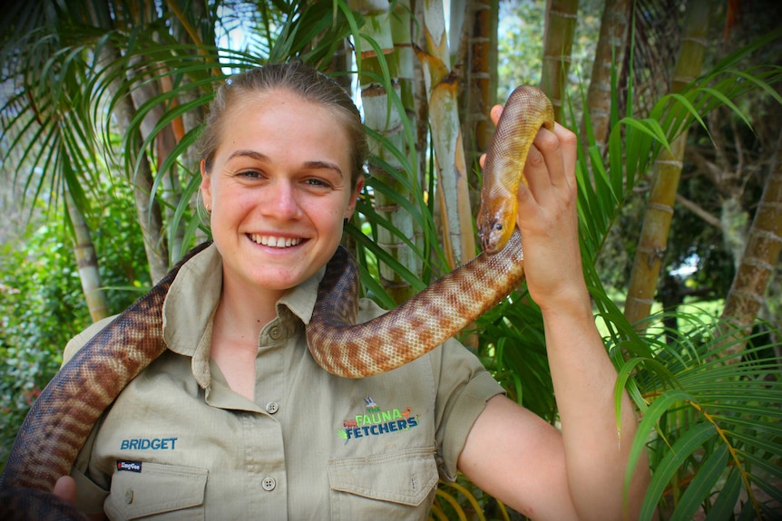 A girl in a khaki shirt holds up a brown and yellow python