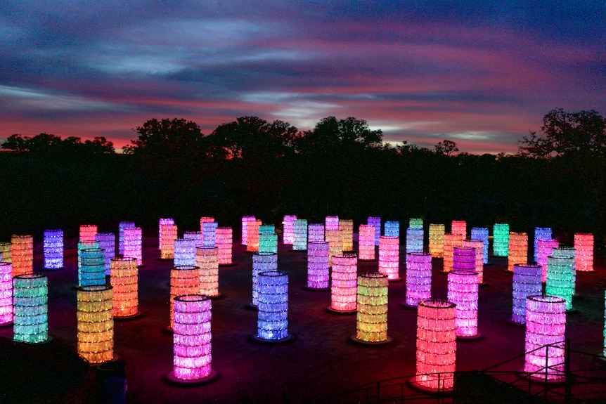 An installation of small colourful, lit up towers, on a field at sunset