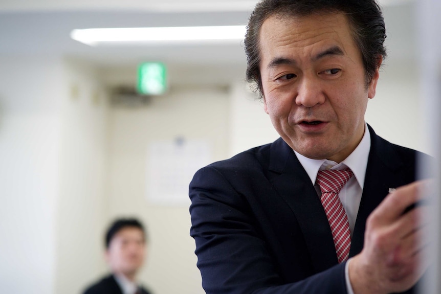 An older Japanese gentleman in a suit writes on a whiteboard