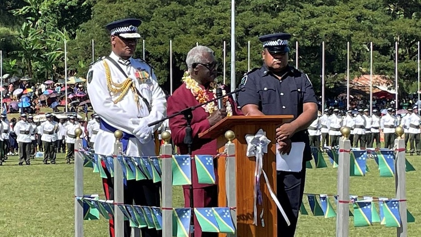 Solomon Islands PM Manasseh Sogavare speaks on a podium wearing a lei and maroon suit flanked by navy, police officers